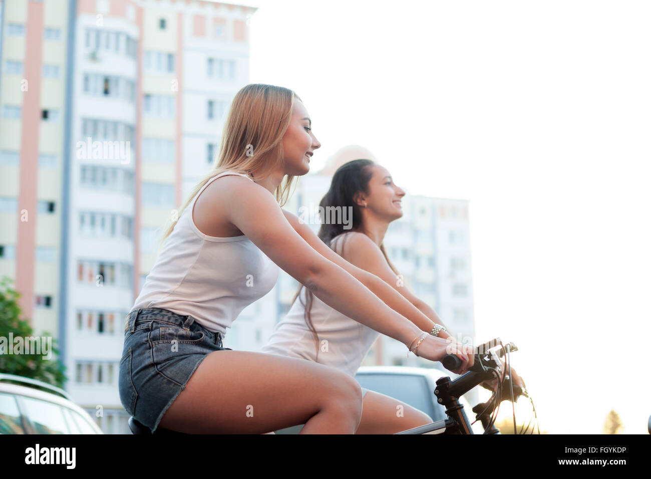 Due sorridenti bella bionda e bruna di donne giovani amici equitazione biciclette in città in estate accanto residential Foto Stock