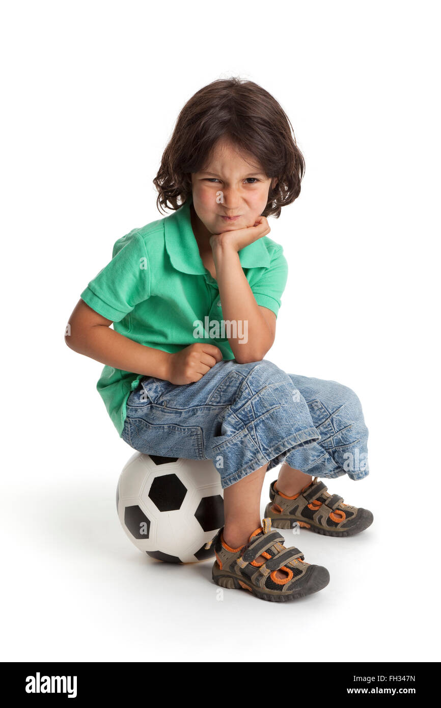 Deluso piccolo ragazzo seduto su un pallone da calcio su sfondo bianco Foto Stock