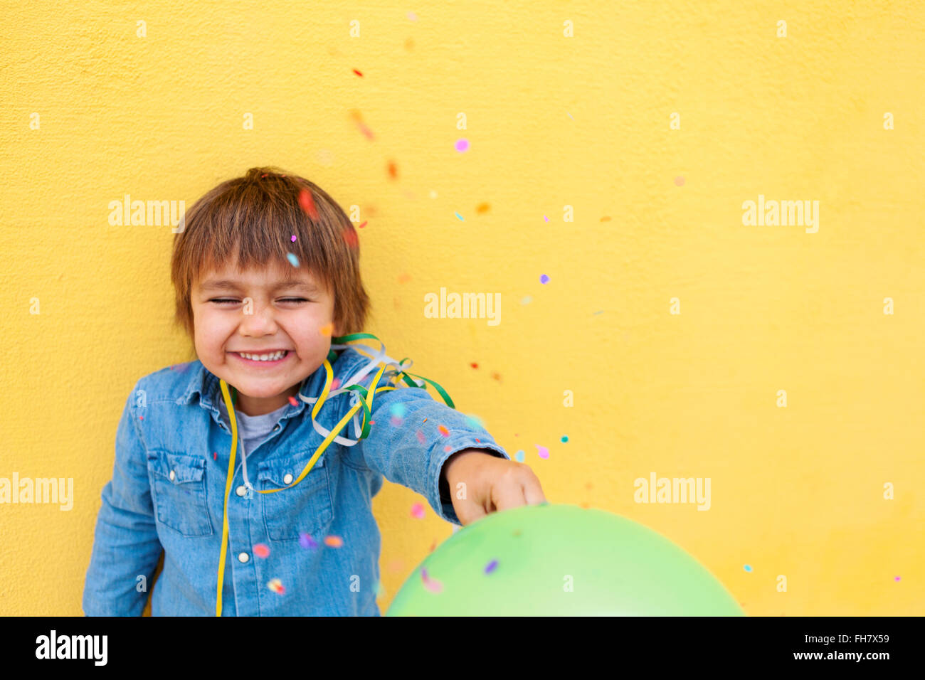 Sorridente ragazzino con palloncino verde, streamer in piedi nella parte anteriore della parete di colore giallo mentre i confetti la caduta verso il basso Foto Stock