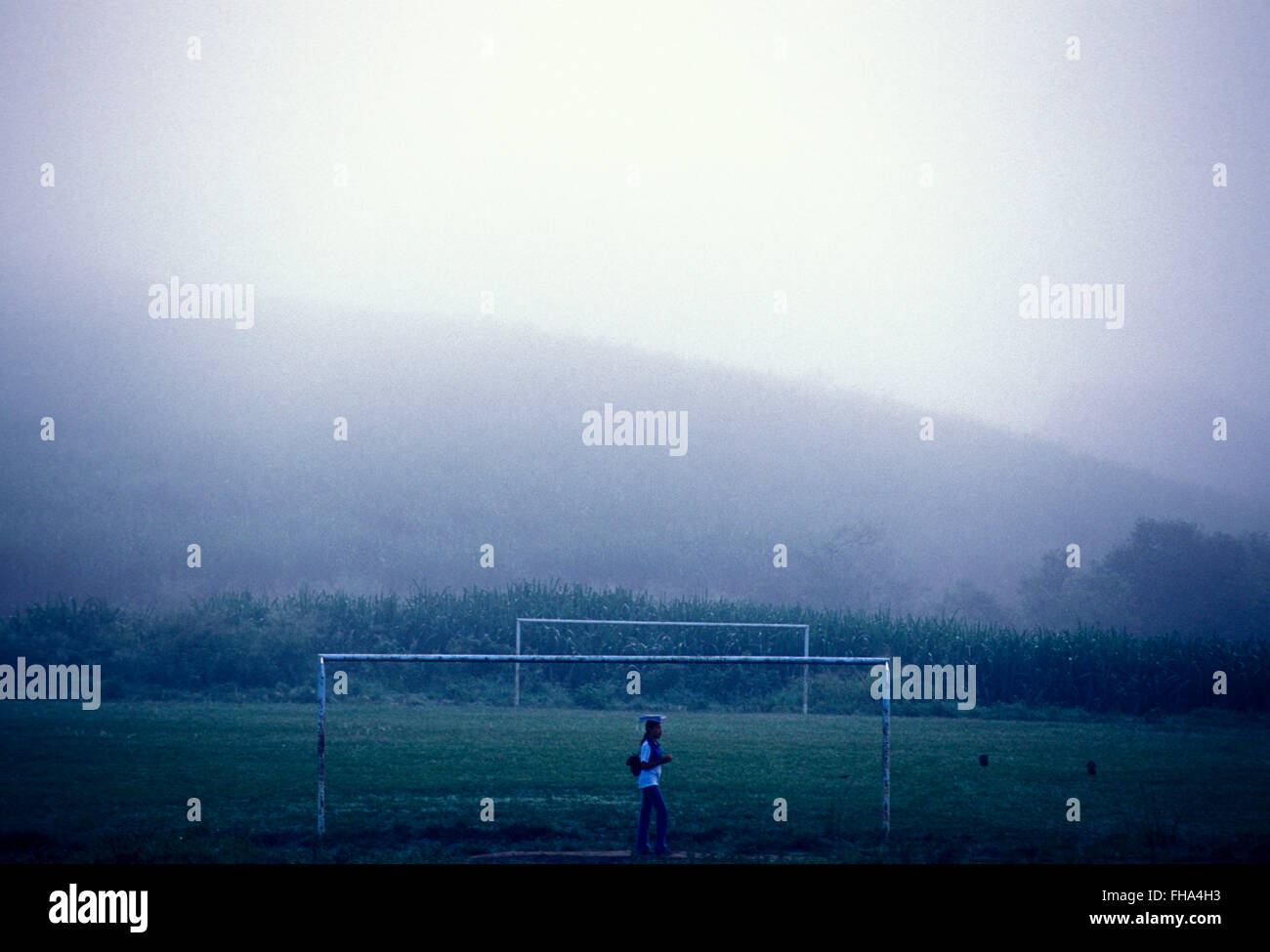 Studente va a scuola rurale di mattina presto e attraversa un campo di calcio vicino alla piantagione di canna da zucchero sotto la nebbia nella zona rurale di Palmares città in stato di Pernambuco, Brasile. Foto Stock