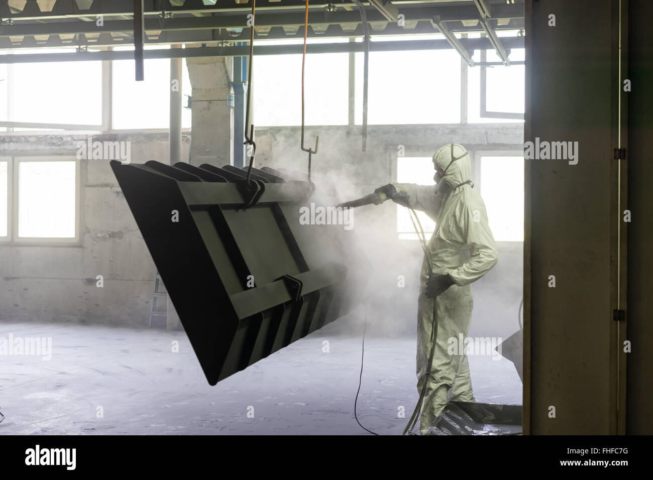 Vista di un lavoratore che indossa una completa bianca tuta protettiva e una maschera per la respirazione, la sabbiatura a metallo cassa appesa a un fascio di metallo in Foto Stock