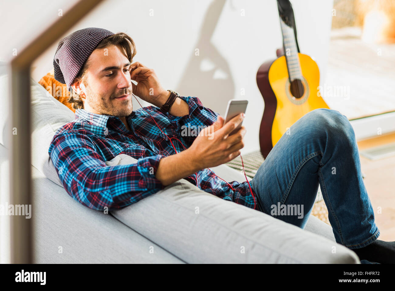 Ritratto di giovane uomo seduto sul divano ascoltando la musica con gli auricolari e lo smartphone Foto Stock