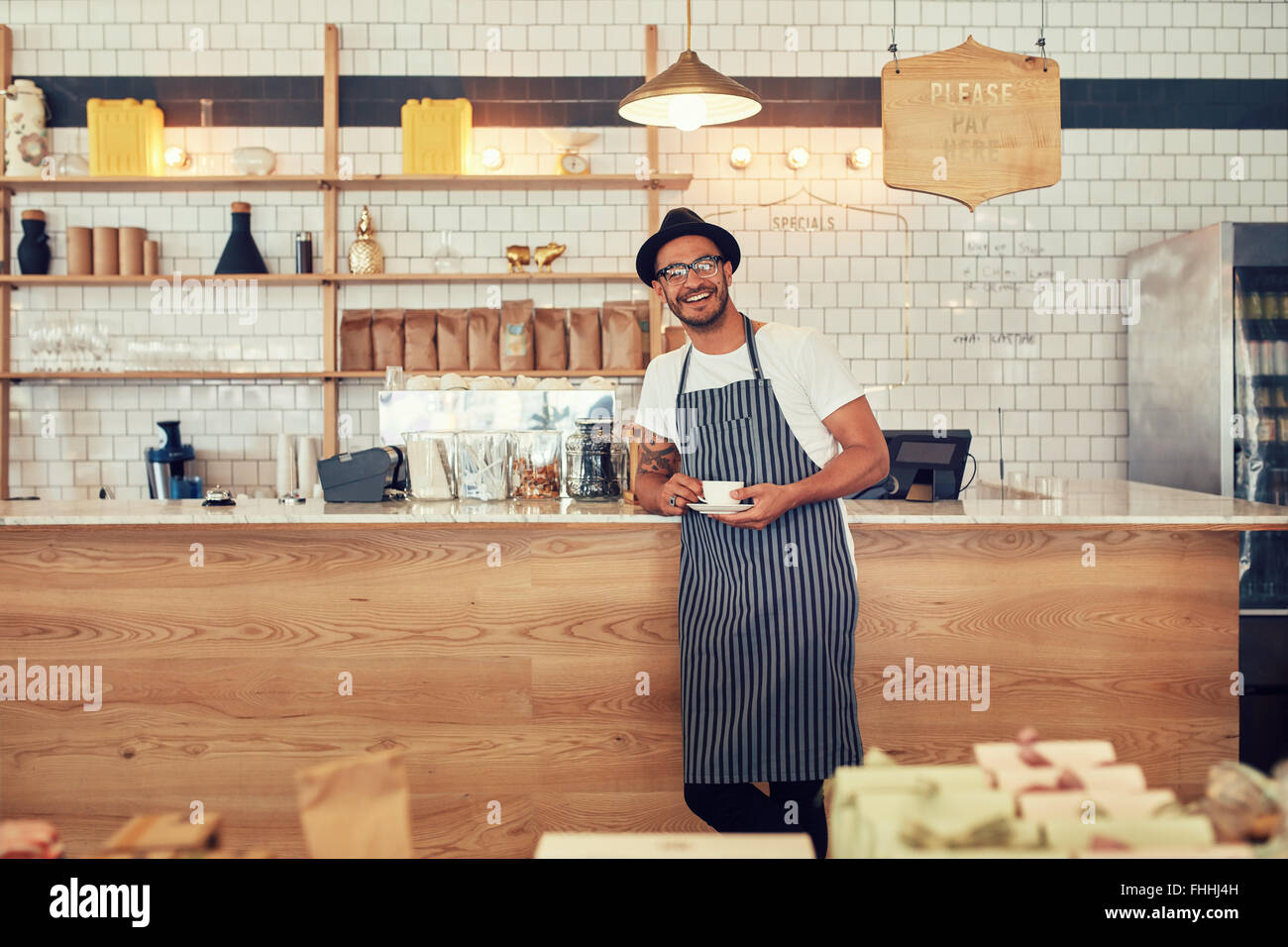 Ritratto di felice giovane uomo che indossa un grembiule e cappello in piedi in un contatore cafe tenendo una tazza di caffè. Coffee shop proprietario looki Foto Stock