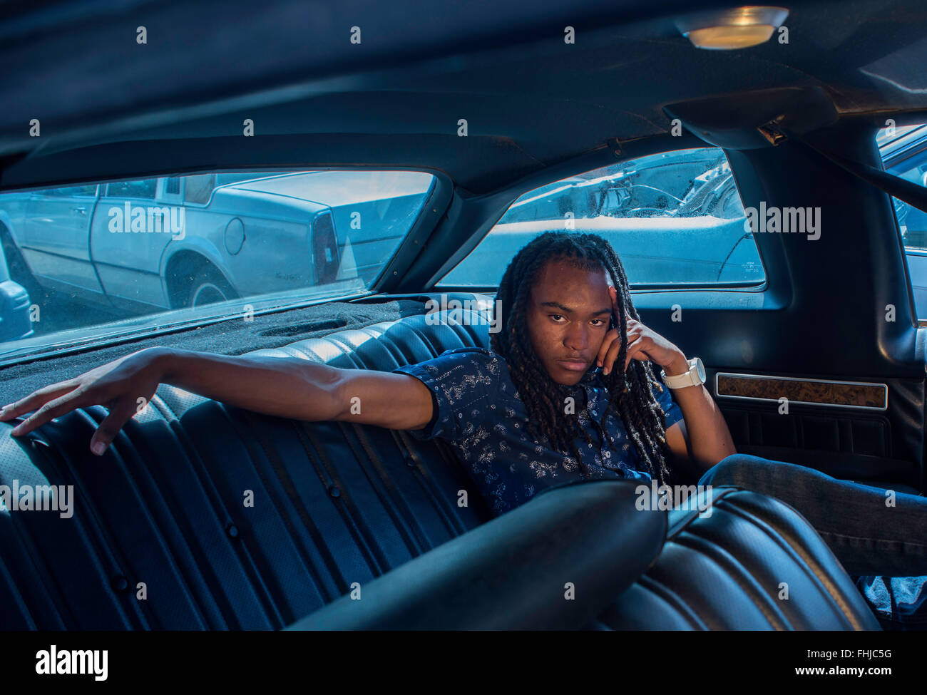 Un afro-americano di adolescente con dreadlocks si erge contro una parete di stagno/siede nel sedile posteriore di un Impala in a junkyard in NC. Foto Stock