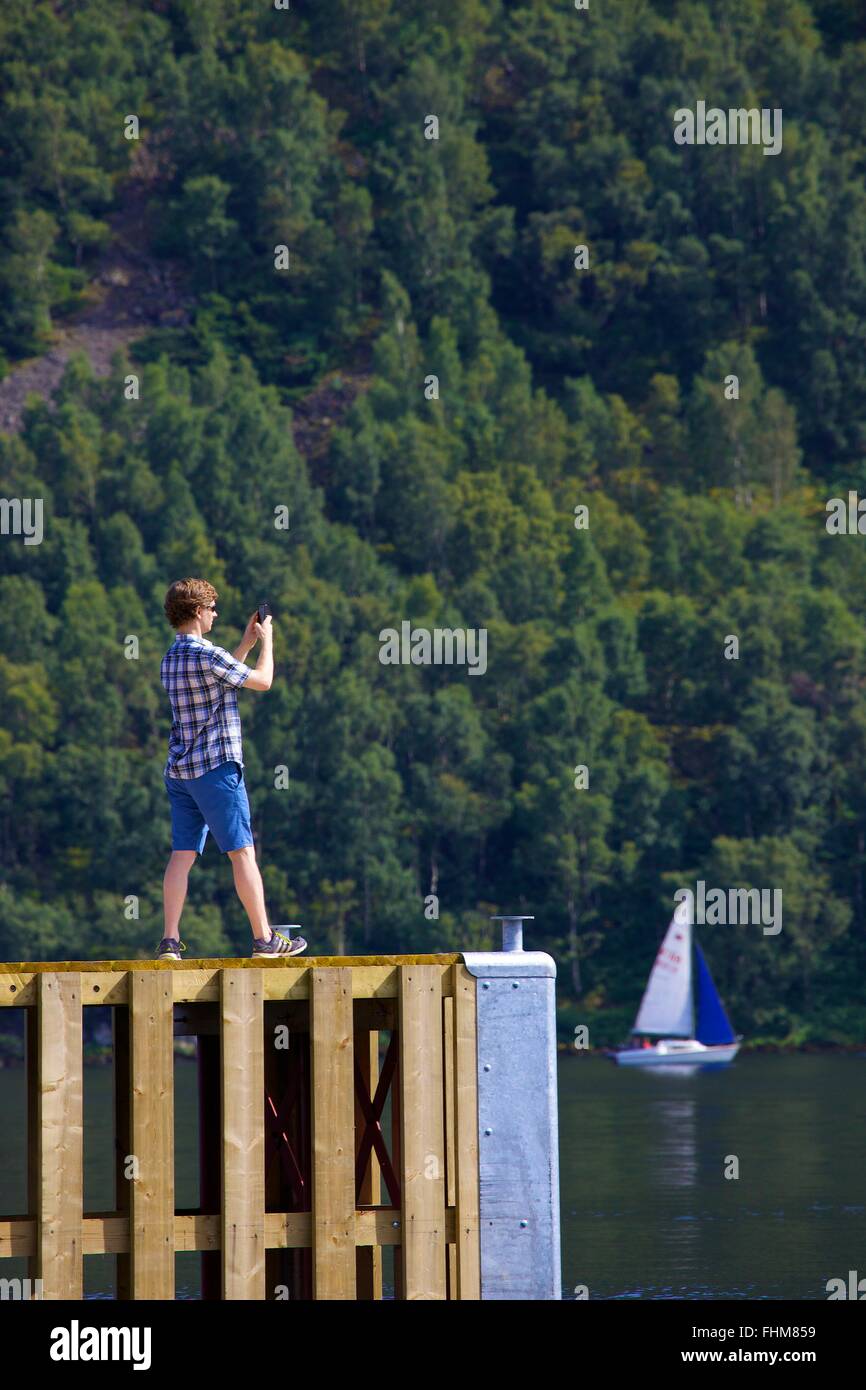 Parco Nazionale del Distretto dei Laghi. L'uomo prendendo fotografie con smart phone da un pontile sul lago Ullswater. Foto Stock