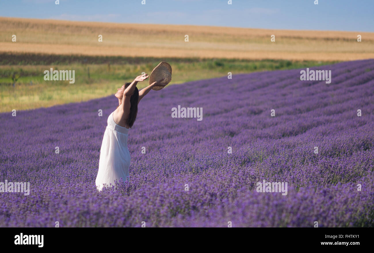 Giovane e bella donna che indossa un abito bianco che celebra la bellezza della vita in piedi al centro di un campo di lavanda in fiore. Foto Stock