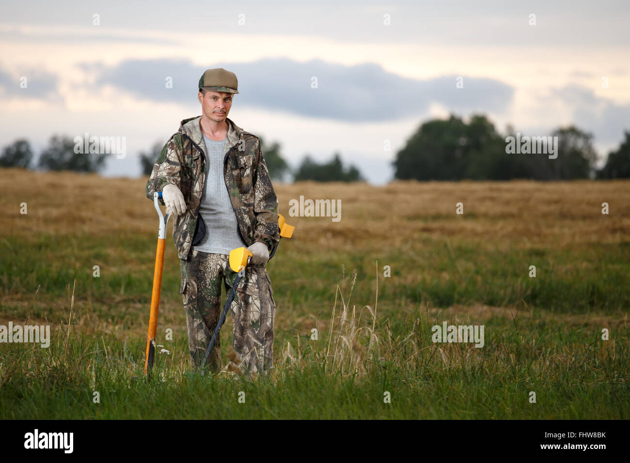 Il cacciatore di tesori con il rivelatore di metalli appoggiato su di una pala in campo. Aspetto fiducioso e camuffare i vestiti. Rurale di fondo Foto Stock