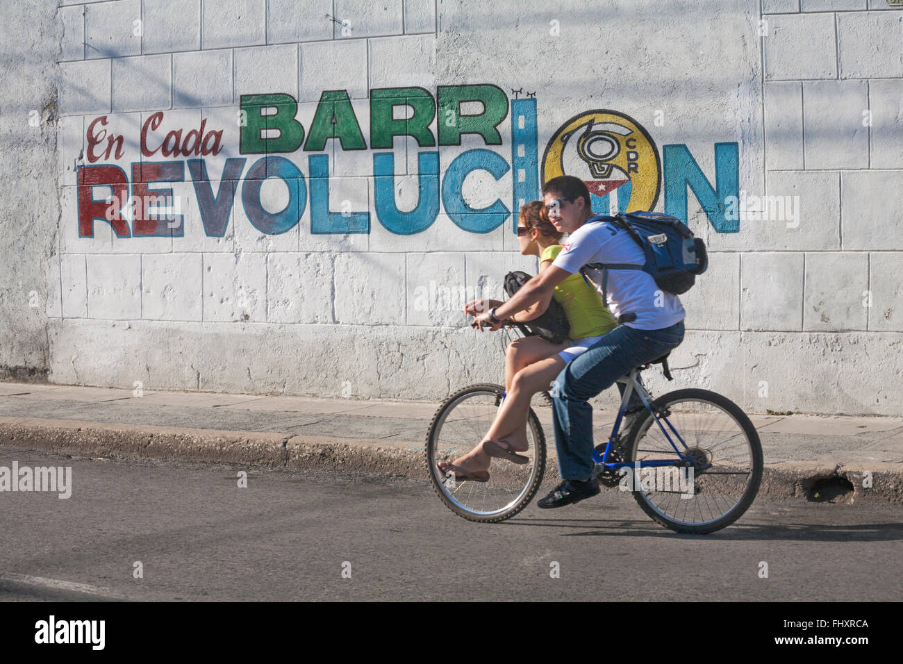 La vita quotidiana a Cuba - Coppia giovane equitazione Bicicletta oltre la parete con en cada barr revolucion slogan a Cienfuegos, Cuba Foto Stock