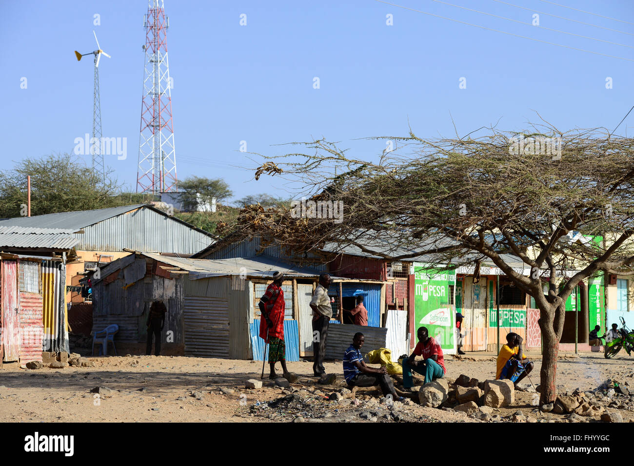 KENYA, Marsabit, villaggio con pilone a traliccio per la telefonia mobile e la trasmissione di piccole windturbine, youngster infront di M-pesa telefono cellulare trasferimento di contanti shop / KENIA, Marsabit, Dorf mit Sendemast fuer Mobilfunk und Kleinwindanlage, herum sitzende Jugendliche vor M-PESA telefono cellulare trasferimento di contanti shop Foto Stock