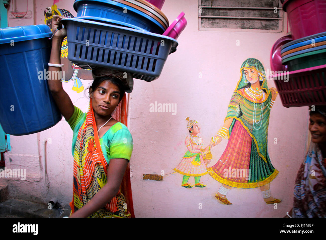 La donna a piedi in una strada di Udaipur, cestello di trasporto sul suo capo, la pittura di una donna con bambino sulla parete, Rajasthan, India Foto Stock