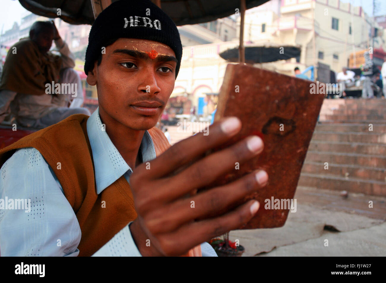 Uomo in Varanasi guardando lo specchio con tilak e indossando 'hero' cap in Varanasi Foto Stock