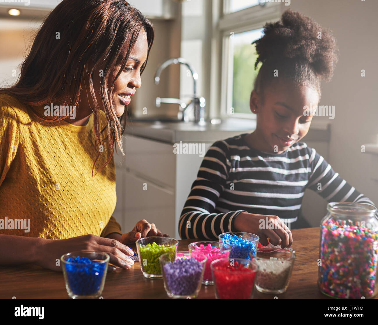 Felice madre africana e figlia creazione di artigianato bordati con diversi colori su un tavolo di legno in cucina con la luce del sole proveniente in Foto Stock