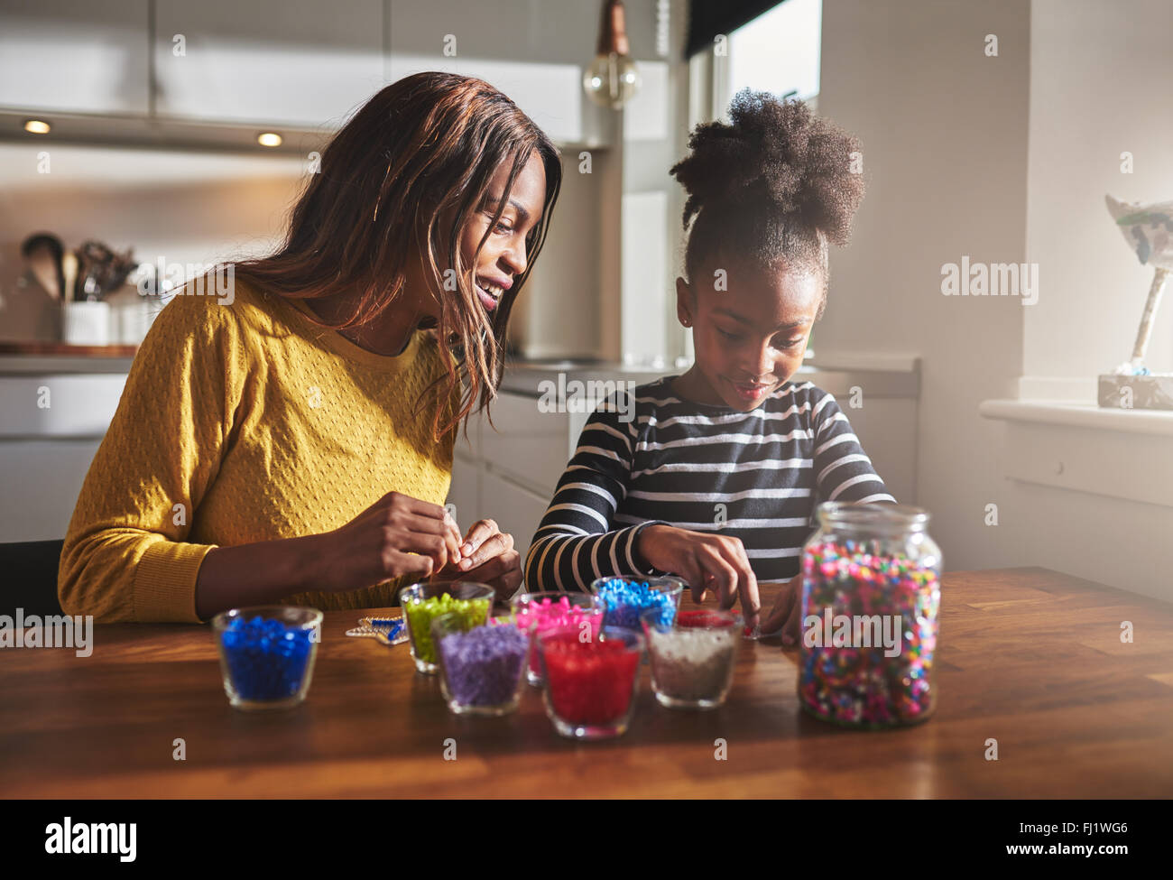 Donna felice e giovane ragazza seduta e la creazione di artigianato bordati con diversi colori su un tavolo di legno in cucina con luce solare comin Foto Stock