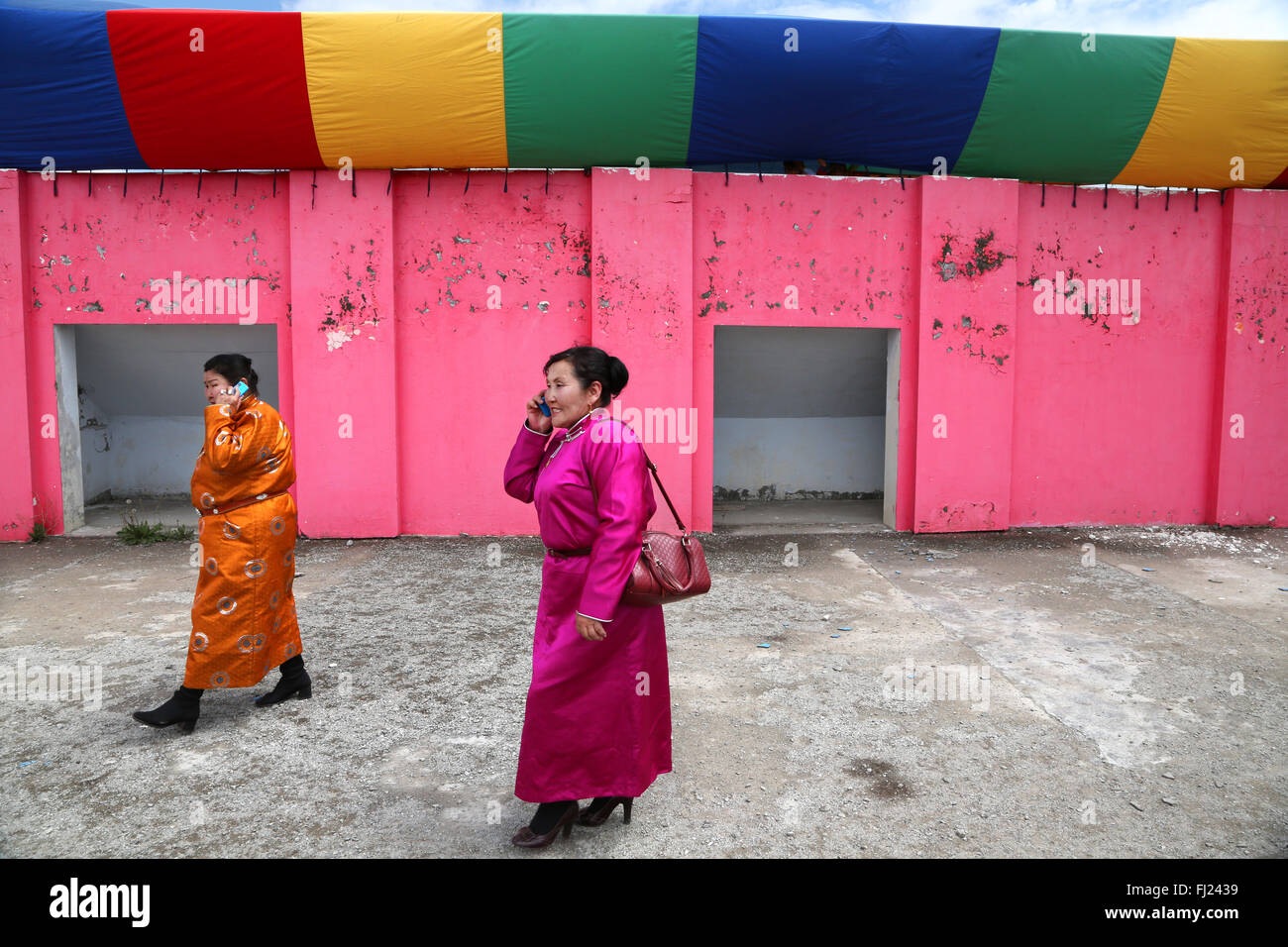 Il mongolo donne con abito tradizionale chiamato 'danguilla a piedi lungo la parete di rosa per le strade di Erdenetwith telefono mobile Foto Stock