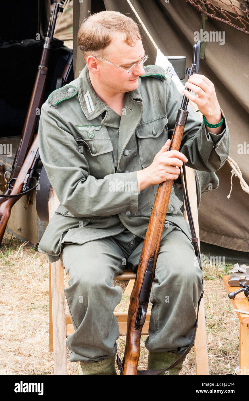 La seconda guerra mondiale la rievocazione. Soldato tedesco seduti su una sedia in esterno ad Encampment, pulizia il suo fucile, tenda dietro di lui. Foto Stock