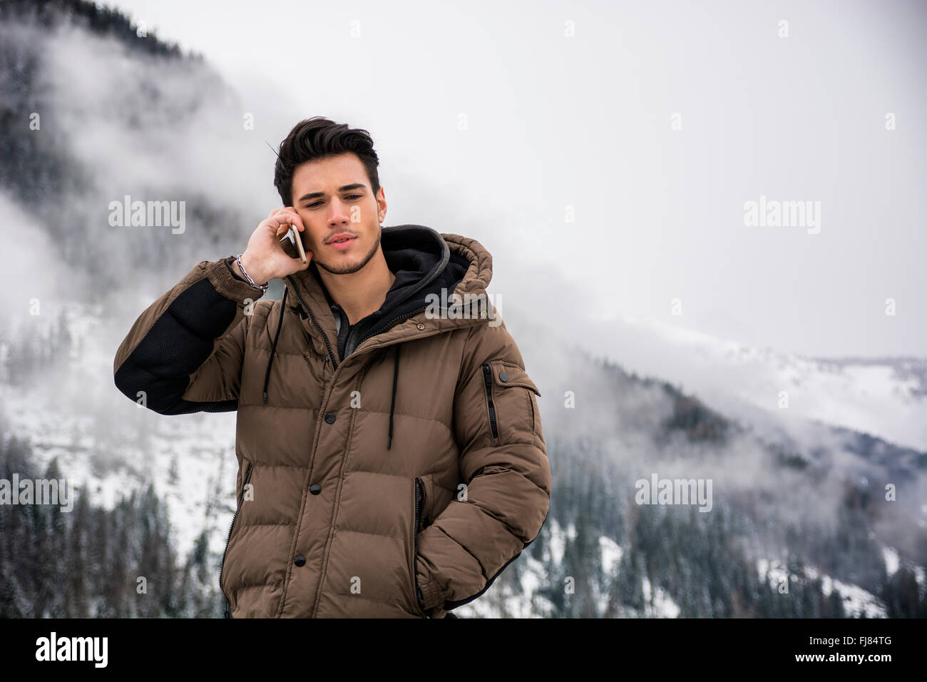 Dai capelli scuri bel giovane in inverno cappotti tramite telefono cellulare o smartphone, outdoor in montagna con paesaggio innevato behi Foto Stock