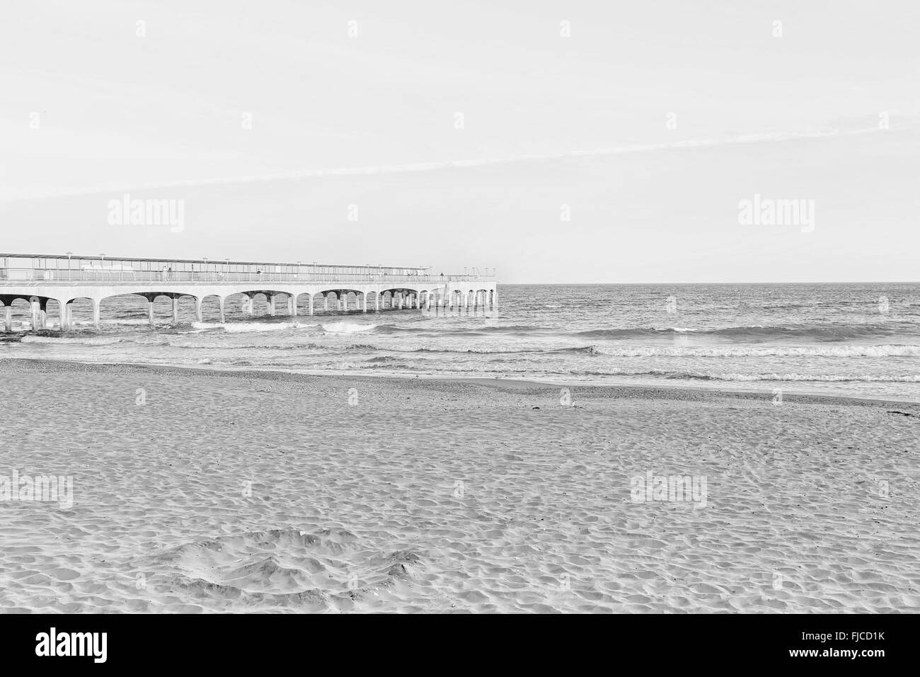 Una immagine in bianco e nero di luce del tramonto di un molo che guarda verso il mare in spiaggia con sabbia e una striscia di drammatiche clo Foto Stock