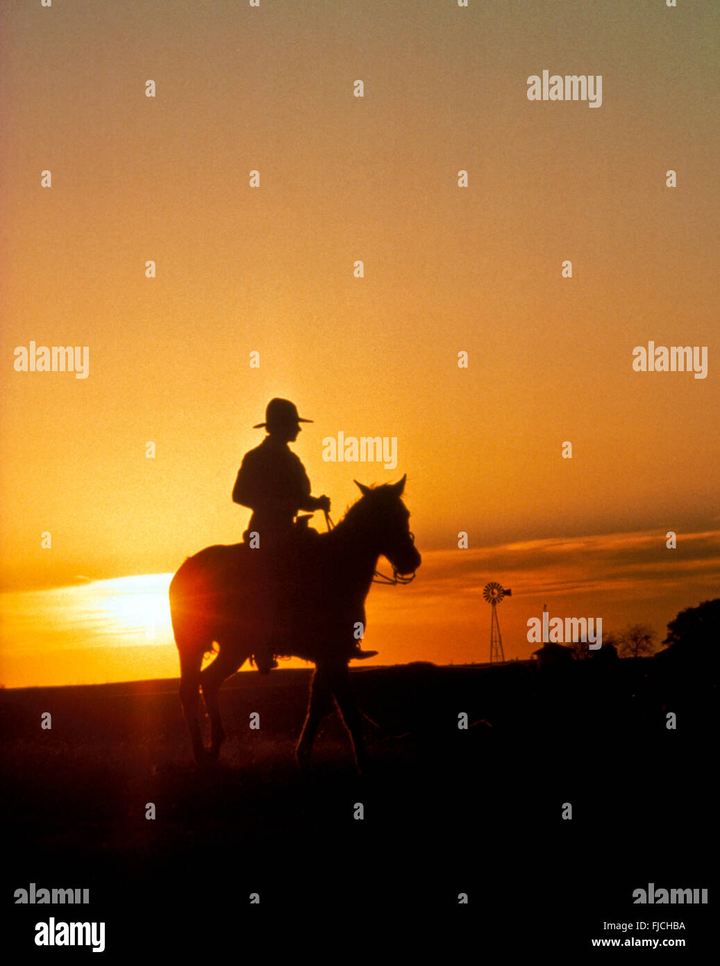 COWBOY AMERICANO, Cowboy in sella al suo cavallo al tramonto con la fattoria e il mulino a vento in background. Idaho, Stati Uniti d'America Foto Stock