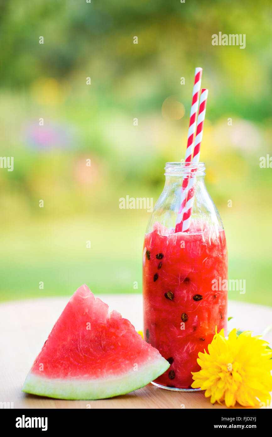 Estivo rinfrescante drink di anguria in una bottiglia con cannuccia un pezzo di cocomero e fiore sulla tavola di legno in giardino Foto Stock