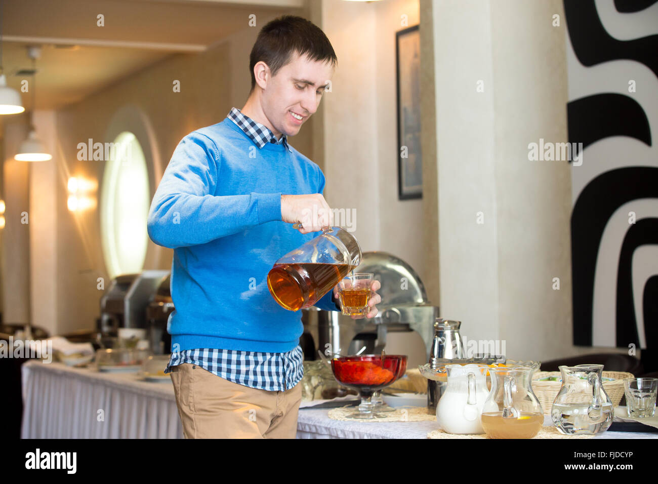 Sorridente casual giovane uomo versando una bevanda dalla brocca in vetro presso il self catering Prima colazione in hotel ristorante al chiuso Foto Stock
