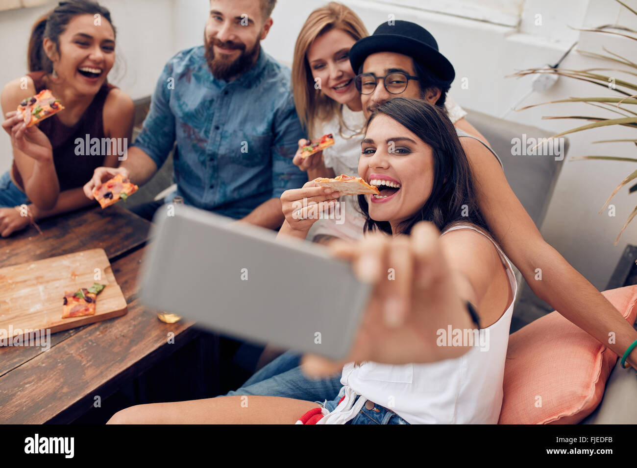 Gruppo di multirazziale giovani prendendo un selfie mentre mangiare la pizza. Giovane donna di mangiare la pizza ai suoi amici seduti intorno durante Foto Stock
