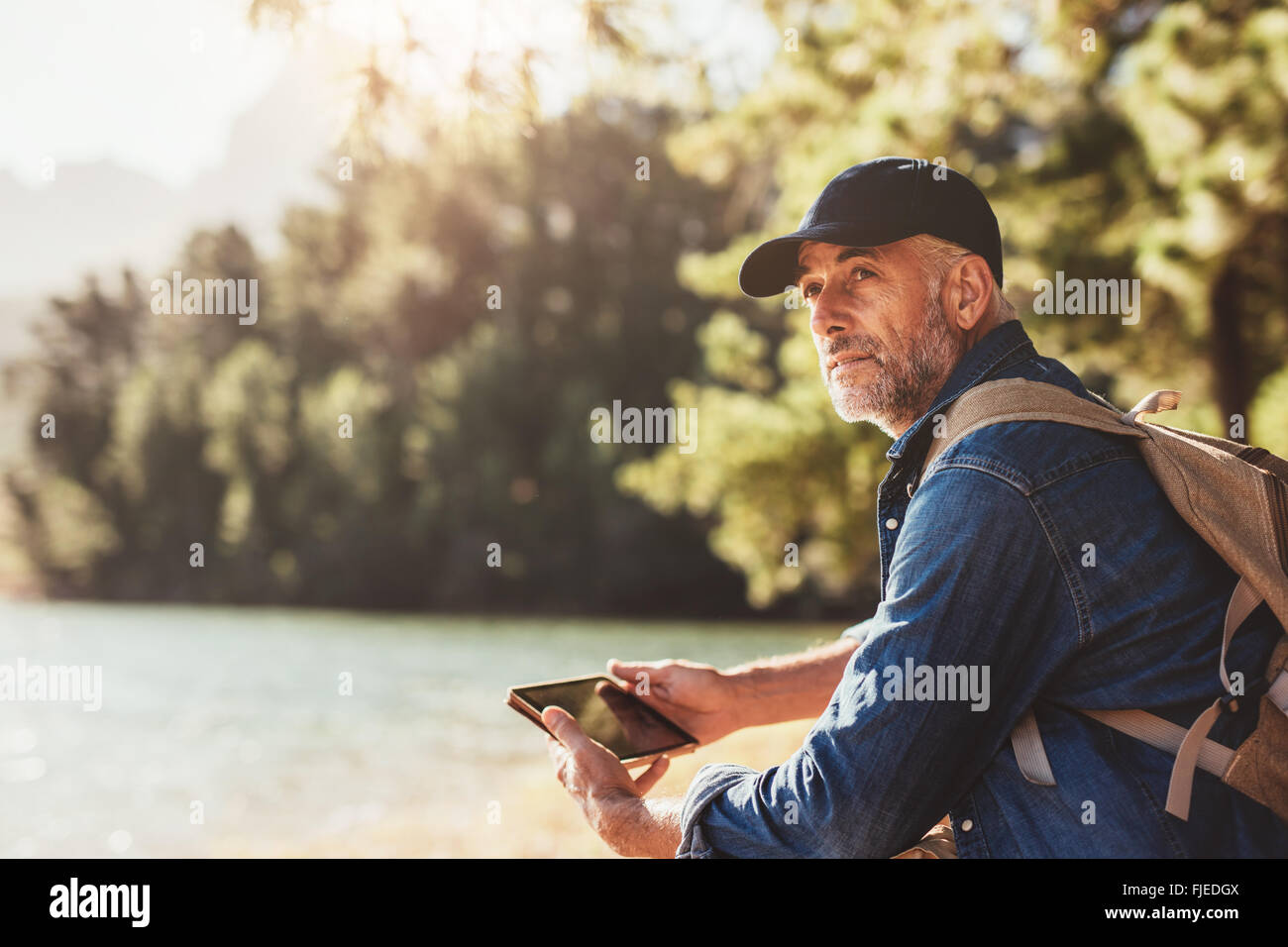 Ritratto di senior uomo seduto vicino a un lago con zaino e digitale compressa. Maschio maturo escursionista seduto di fronte ad un lago e cercando di awa Foto Stock