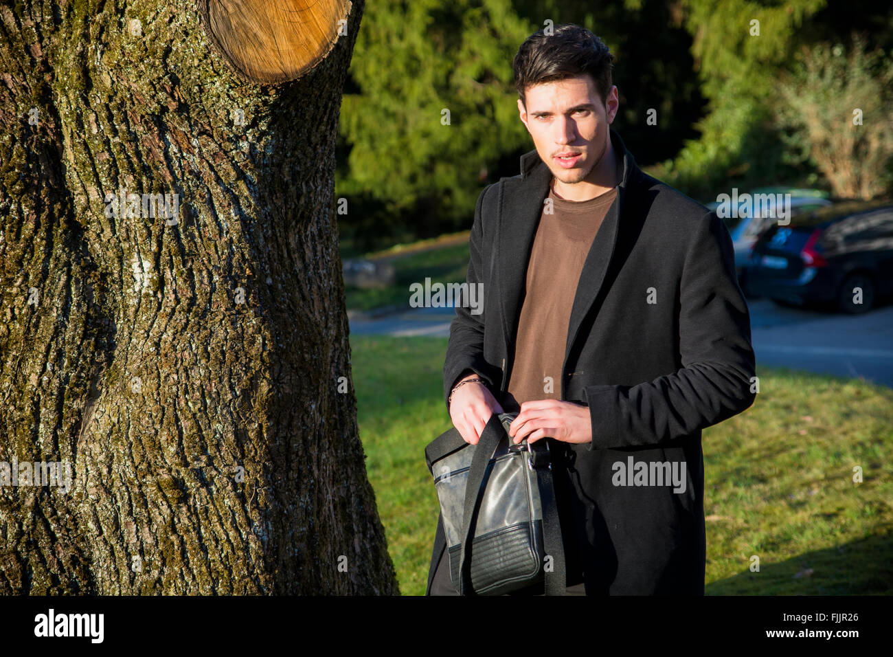 Bel giovane uomo appoggiato contro l'albero, guardando la telecamera, in una giornata di sole che indossa un mantello nero Foto Stock