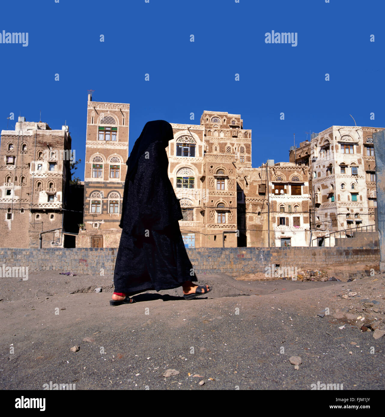 Una donna musulmana velata passeggiate su una sana di una strada, Yemen.a sfondo tipiche case dello Yemen. Foto Stock