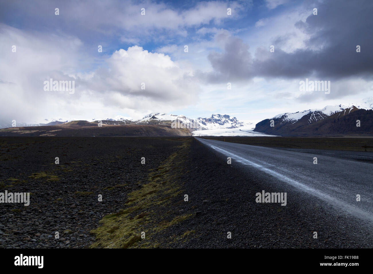 Foto orizzontale di una dritta strada asfaltata proveniente dalle montagne con le nuvole sopra e il ghiacciaio Vatnajokull in backg Foto Stock