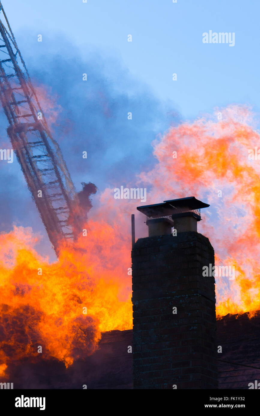 Un lone pompiere travolto in ondate di calore decends una scala nel retro di un fiammante tetto. Foto Stock