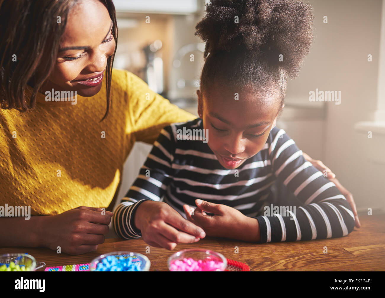 Felice madre in giallo camicetta e bambino lavora con perle di vari colori in vasi su un tavolo di legno Foto Stock