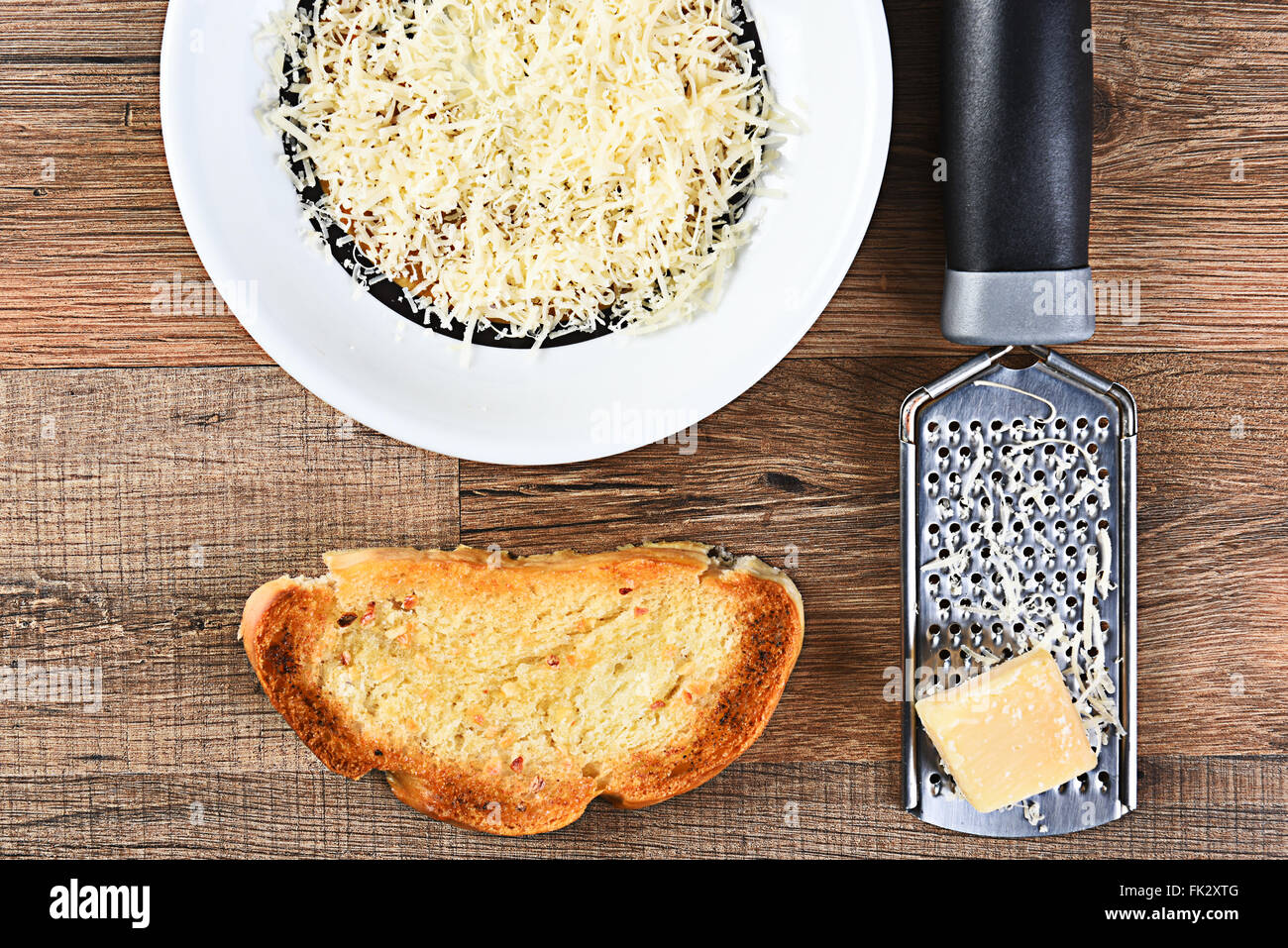 Elevato angolo di visione di un pezzo di pane all'aglio e una ciotola di parmigiano grattugiato con una grattugia e grumo di formaggio. Foto Stock