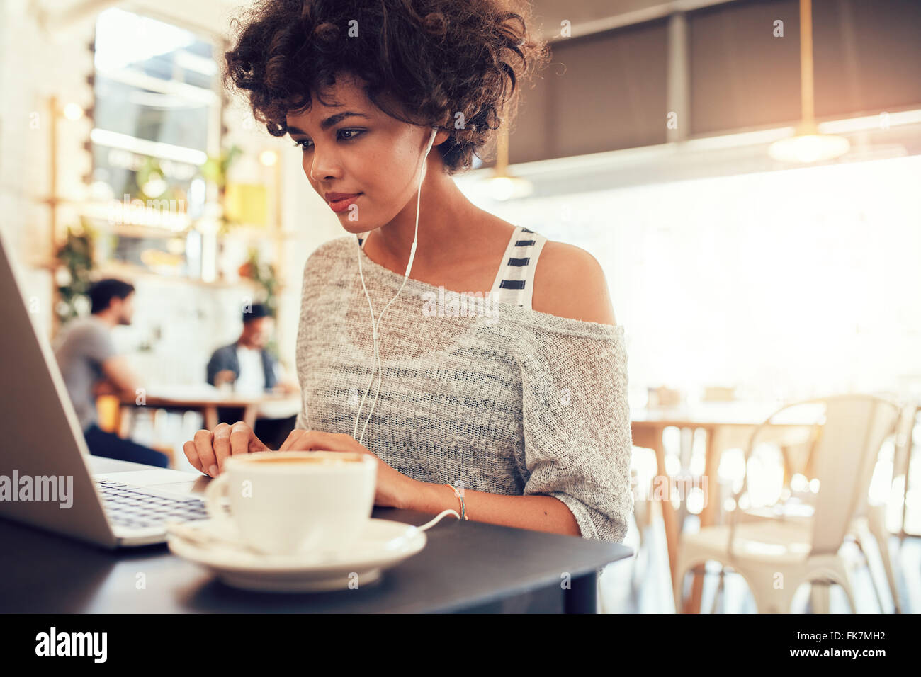 Ritratto di un attraente giovane donna con cuffie utilizzando il portatile in un cafe'. African American donna lavora sul computer portatile di un Foto Stock