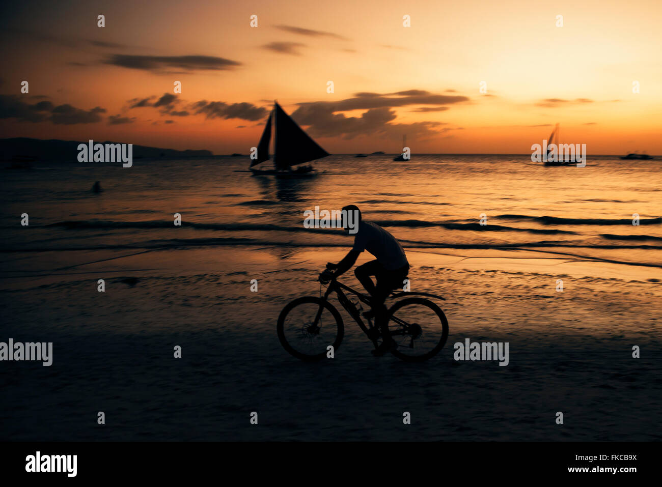 Silhouette di un uomo corse in bicicletta in Boracay beach Filippine. Ci sono barche a vela sagome sul bacgrkound al tramonto. Foto Stock