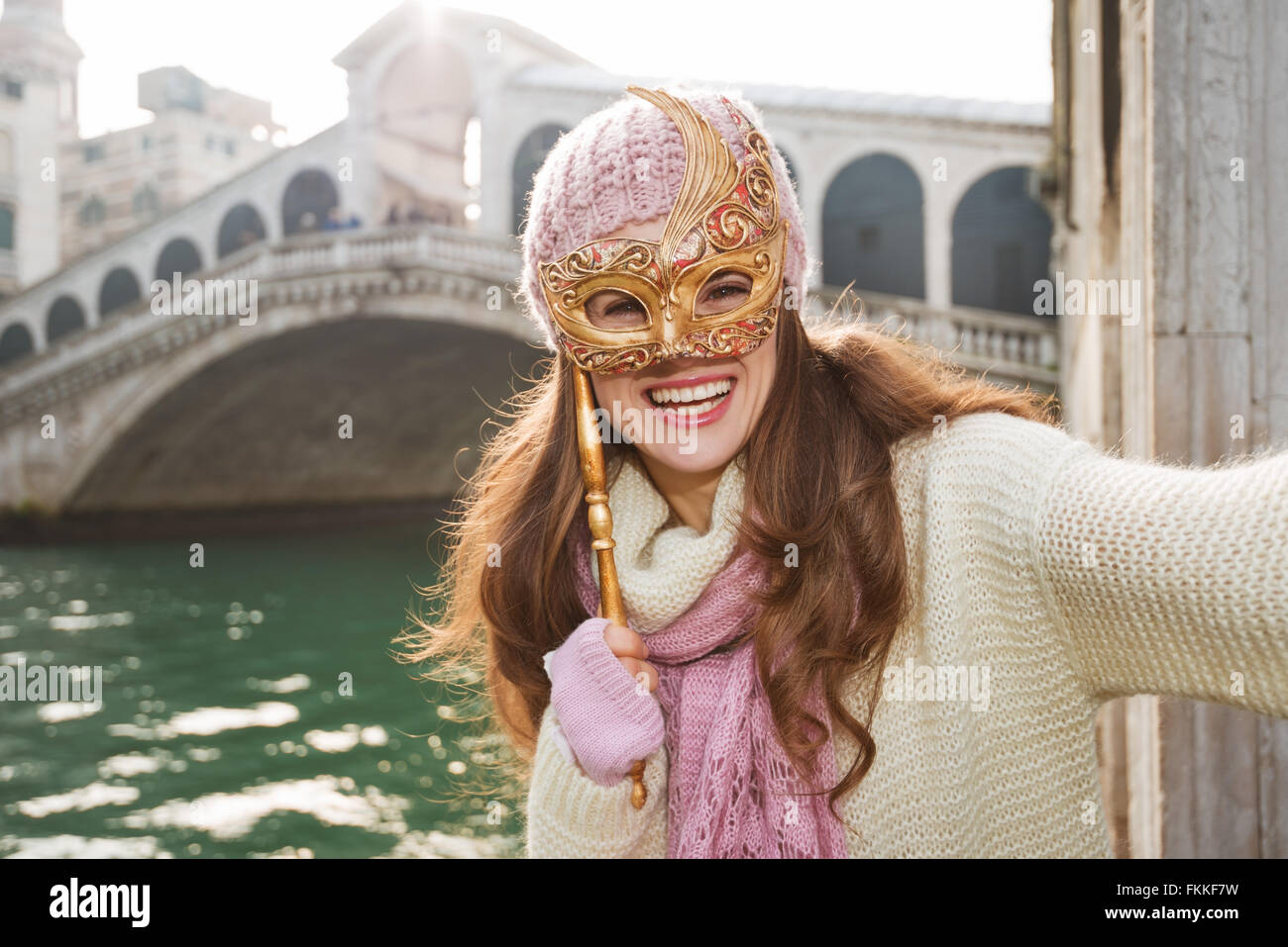 Ultimate getaway collegamento - Inizio Anno Nuovo andando sul Carnevale di Venezia, Italia. Sorridente giovane donna in bianco maglione di nascondersi dietro la maschera di Venezia nella parte anteriore del ponte di Rialto e tenendo selfie Foto Stock