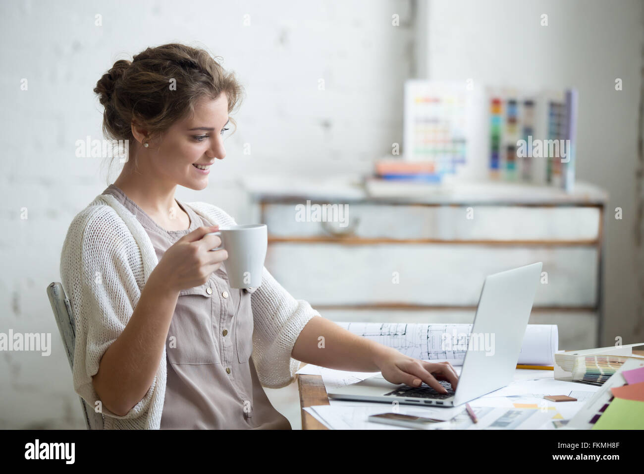 Ritratto di belle sorridenti giovane designer donna seduta a home office desk con tazza di caffè, lavorando sul computer portatile Foto Stock