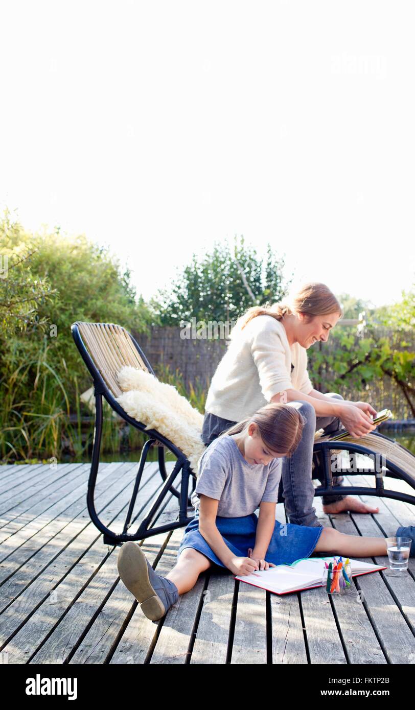 Ragazza disegno su decking con la madre seduta sulla sedia a sdraio Foto Stock