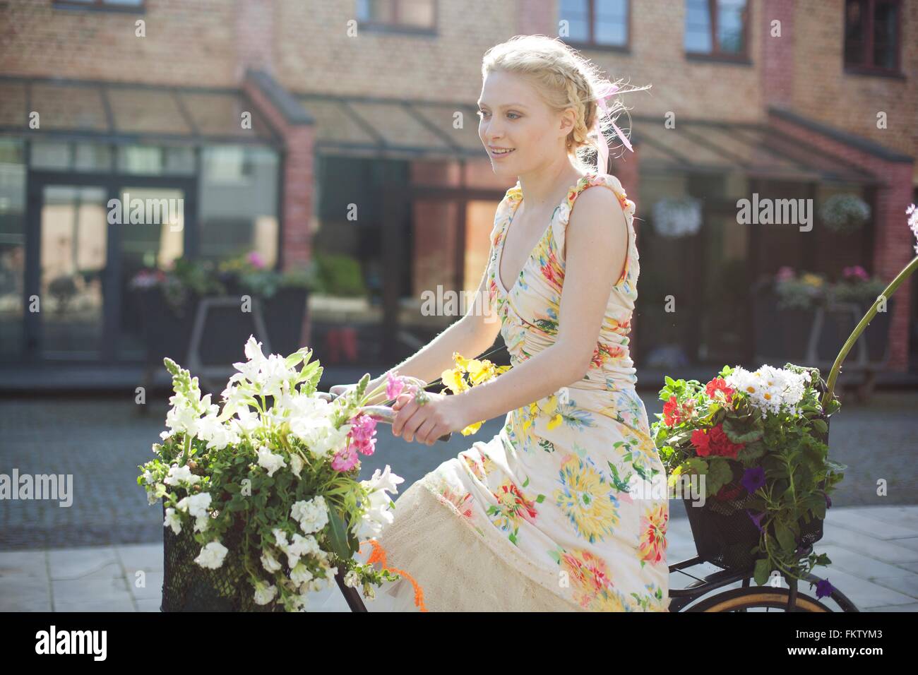 Giovane donna escursioni in bicicletta lungo la strada, che trasportano fiori in cesti in bici Foto Stock