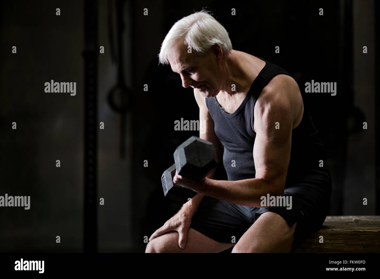 Senior uomo seduto facendo bicipite ricci con il manubrio in palestra scuro Foto Stock