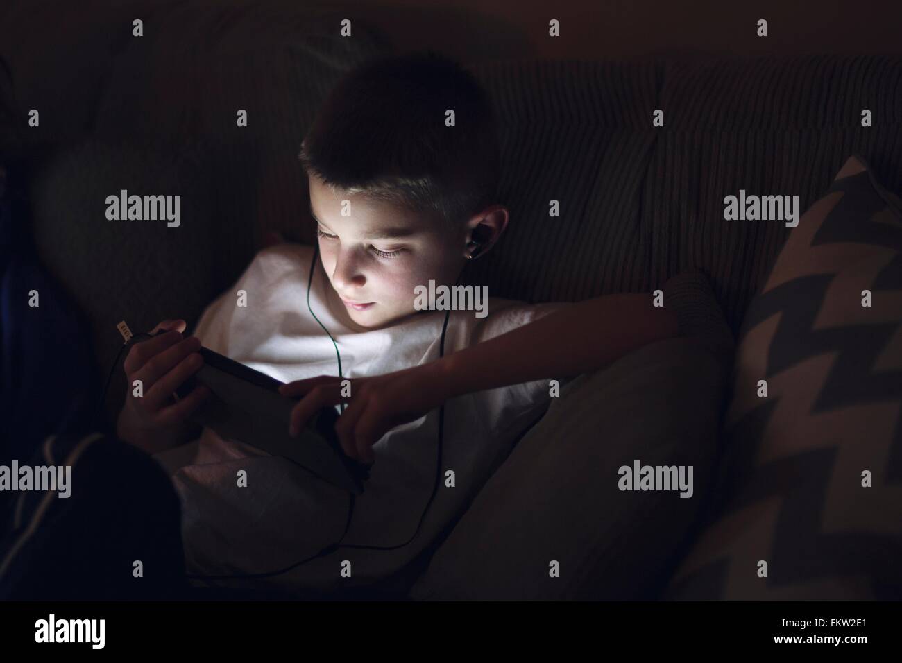 Ragazzo sul divano che indossa gli auricolari guardando verso il basso utilizzando tavoletta digitale Foto Stock