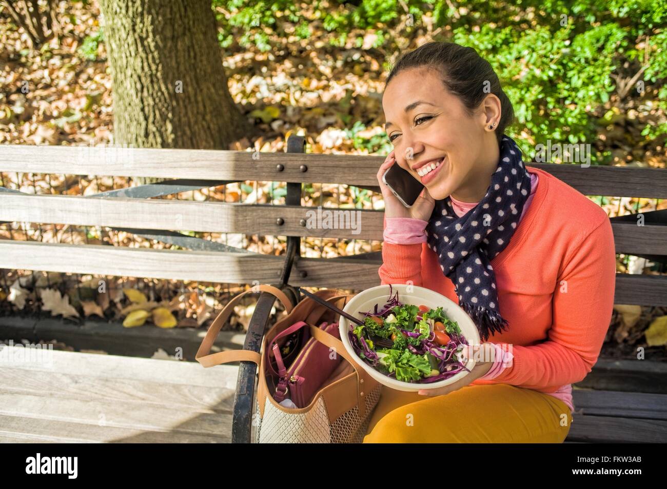 Giovane donna seduta su una panchina nel parco chiacchierando sullo smartphone mentre si mangia il pranzo Foto Stock