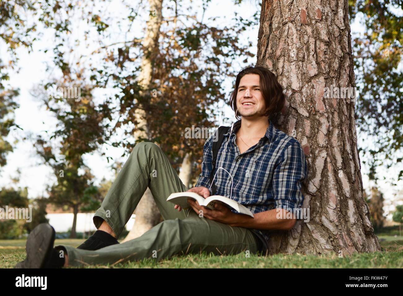 Giovane uomo seduto contro la struttura di libro di contenimento, guardando lontano sorridente Foto Stock