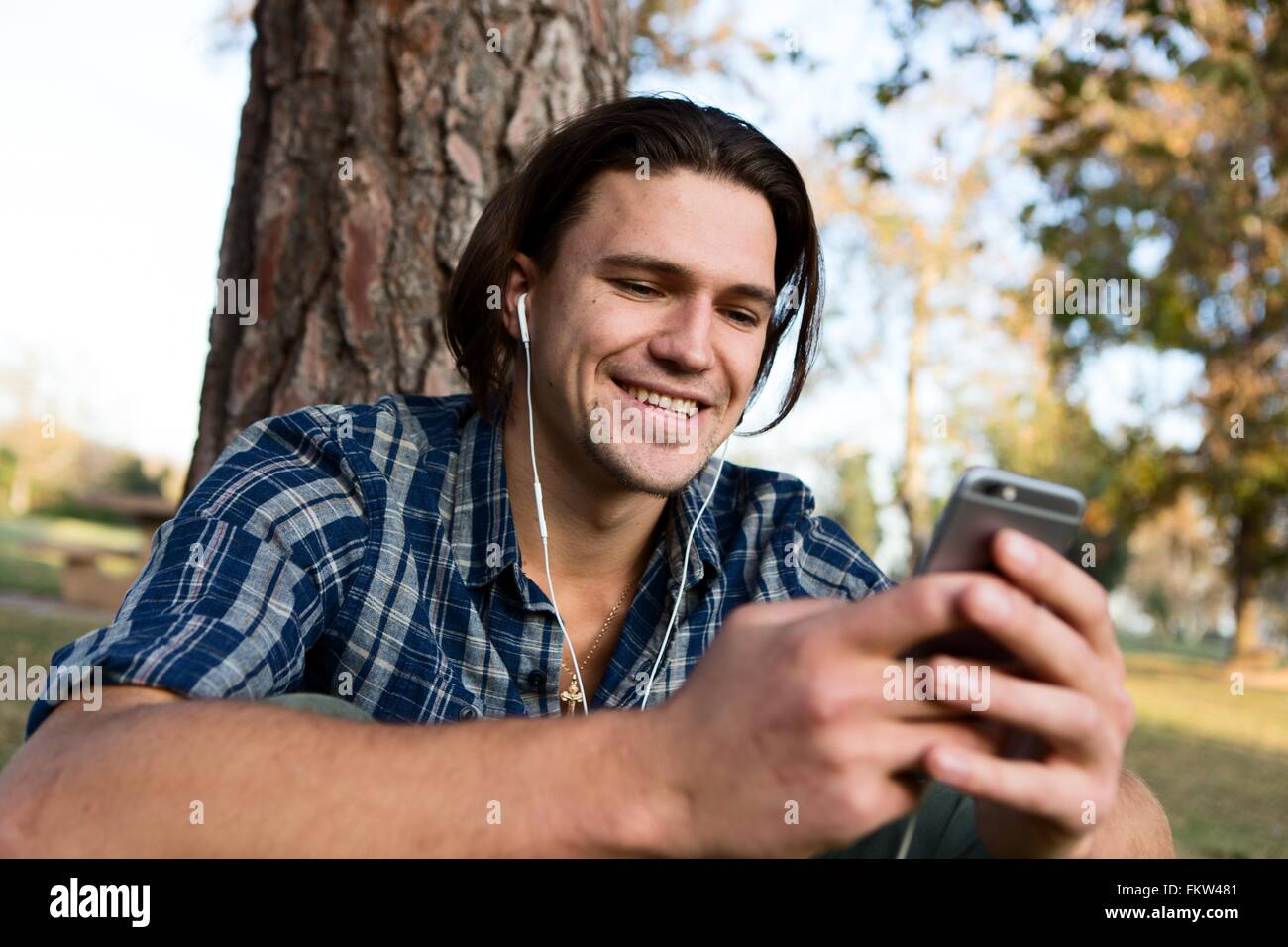 Giovane uomo seduto mediante una struttura ad albero che indossa gli auricolari guardando sorridente dello smartphone Foto Stock