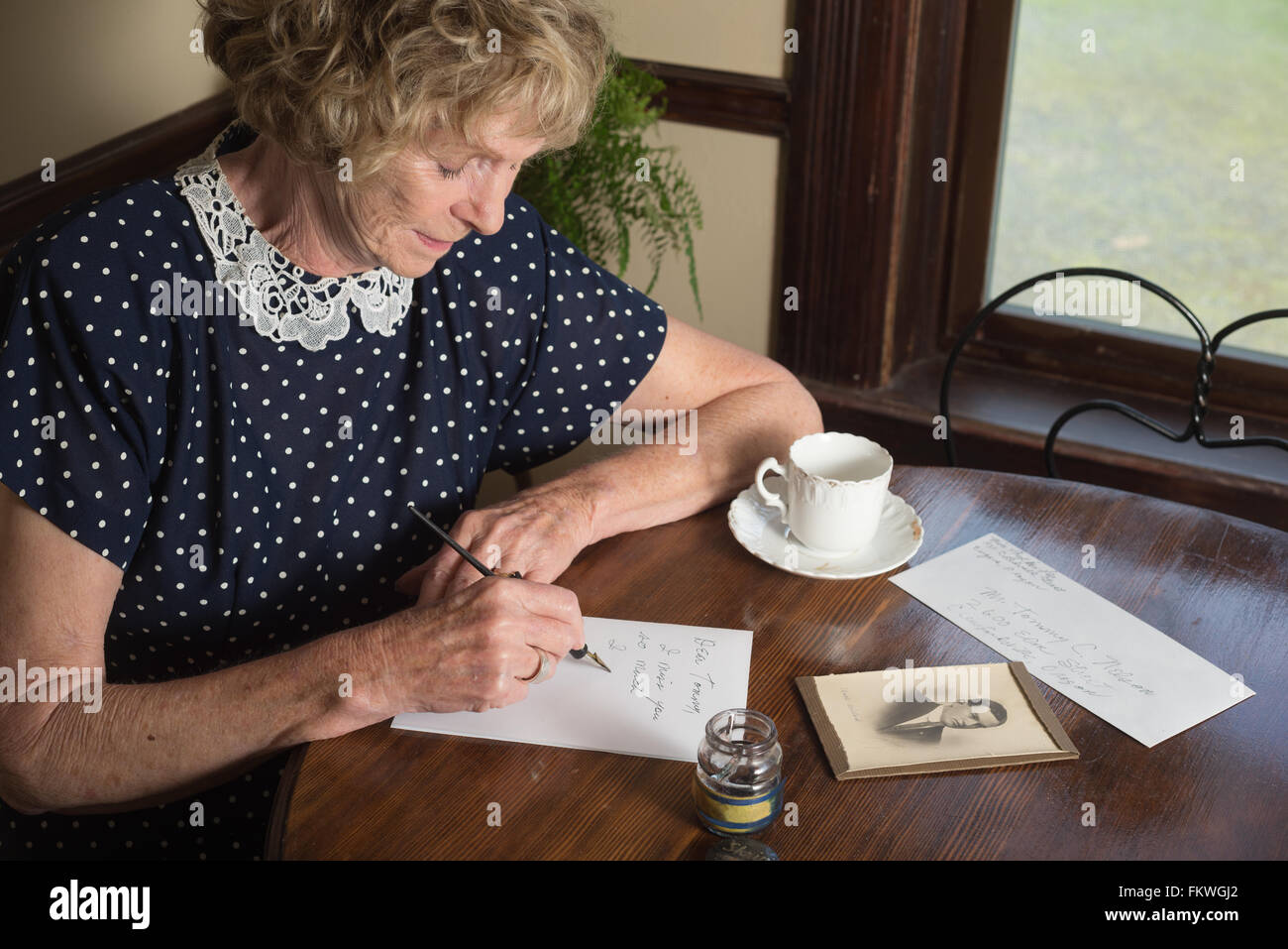 In una scena storica degli anni trenta o quaranta, un attraente donna anziana scrive una lettera come si siede a un tavolo da una finestra. Foto Stock