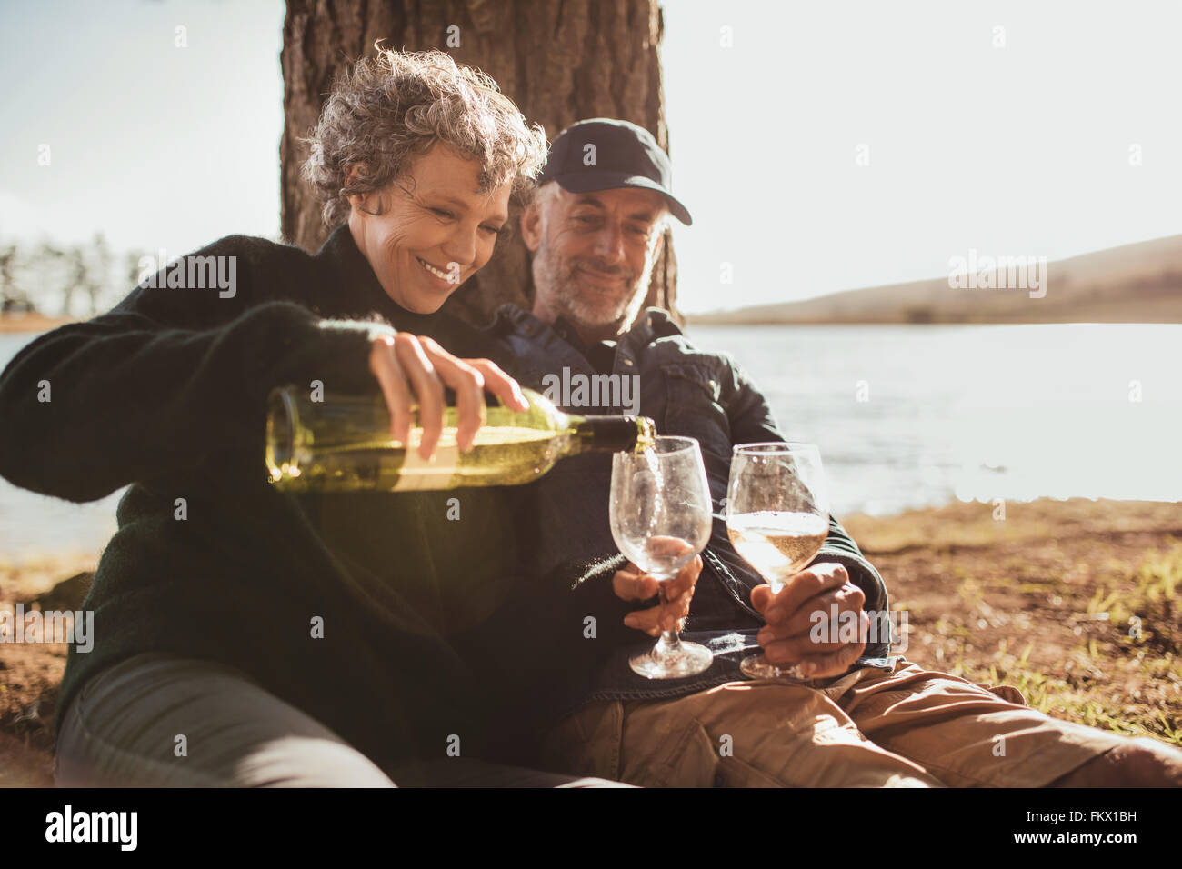 Coppia senior godendo bevande al campeggio vicino al lago. Donna matura versando il vino in bicchieri, sia seduto sotto un albero in un estate Foto Stock