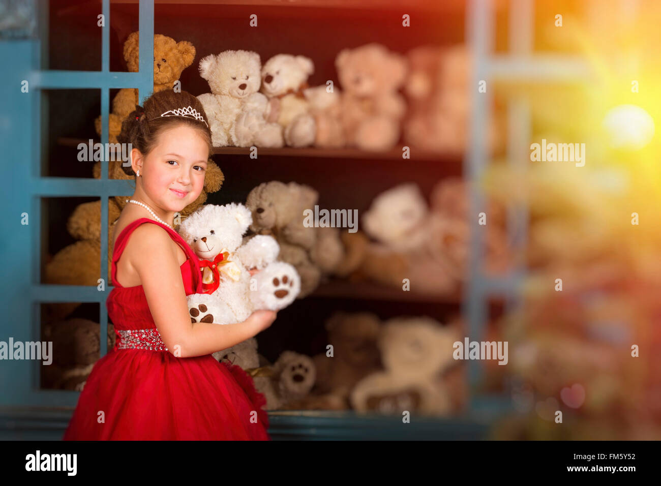 Adorabile bambina vestito in un lussureggiante abito rosso, abbracciando un orsacchiotto di peluche accanto alla finestra con i giocattoli Foto Stock