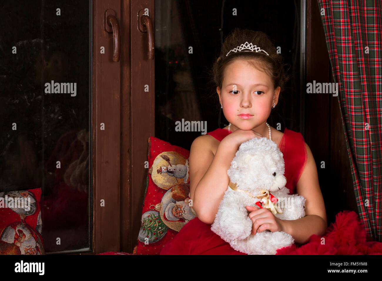 Riflessivo adorabile bambina vestito in un lussureggiante abito rosso, abbracciando un orso di peluche accanto alla finestra Foto Stock