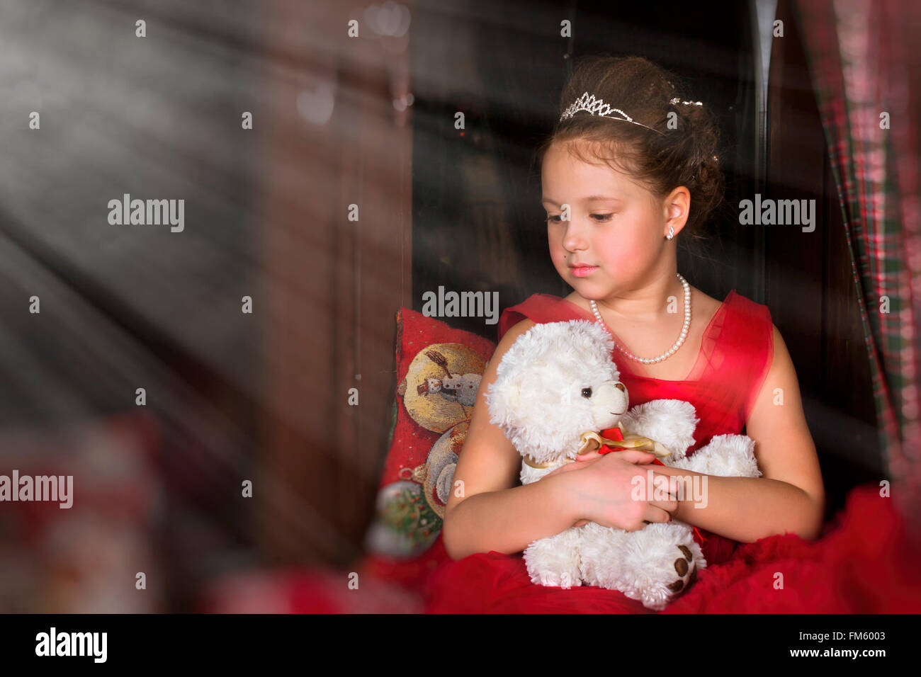 Riflessivo adorabile bambina vestito in un lussureggiante abito rosso, abbracciando un orso di peluche accanto alla finestra Foto Stock