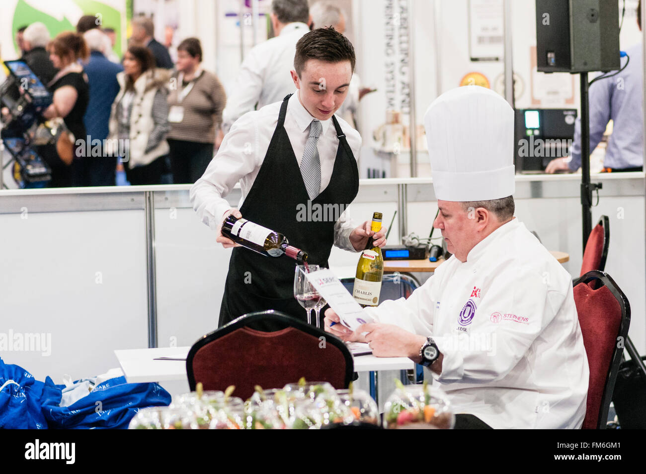 Un cameriere versa un bicchiere di vino rosso per un cuoco, come egli controlla il menu in una competizione in una mostra di catering Foto Stock