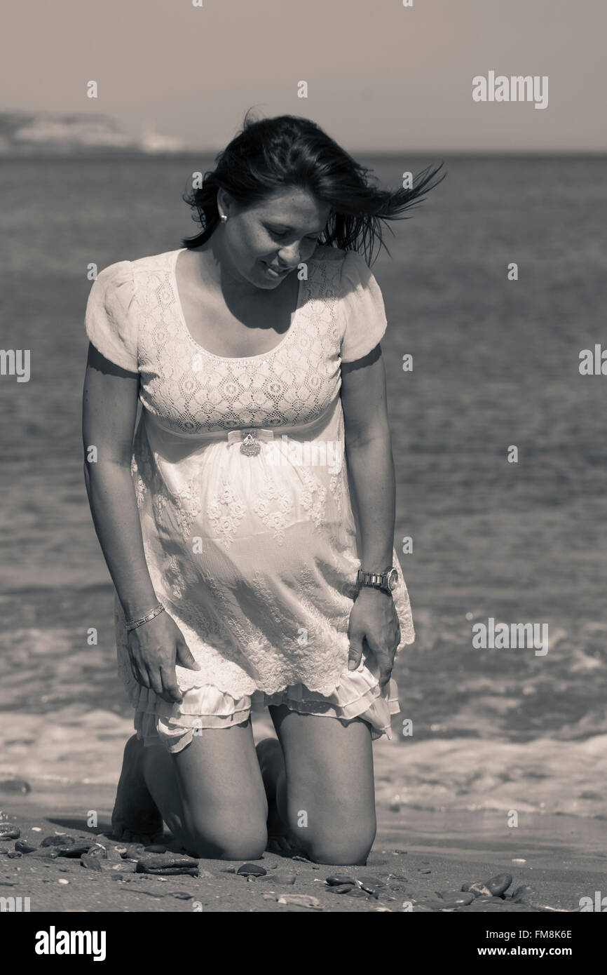 Ritratto di una bella felice donna incinta sulla spiaggia, in ginocchio e guardando verso il basso. Focus sul ventre. Foto Stock
